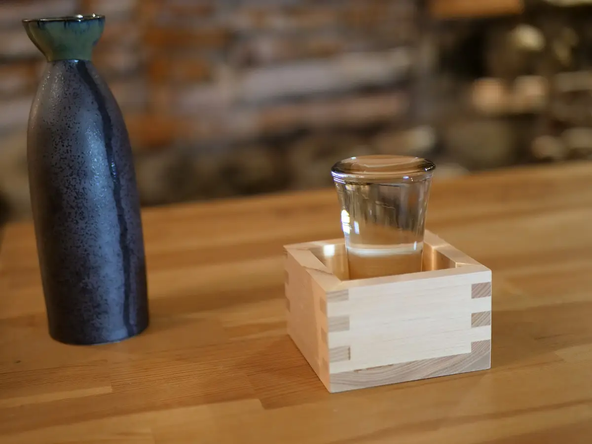 A glass of sake overflowing into a wooden masu box, next to a dark ceramic flask